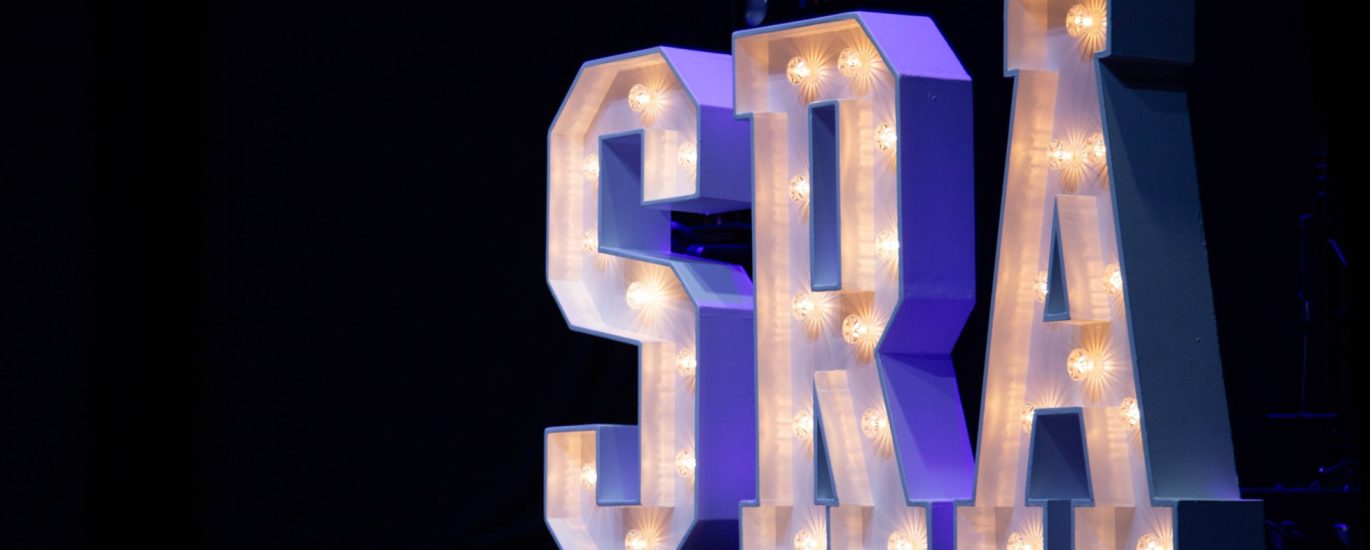 Large illuminated letters spelling S R A, on a stage