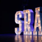 Large illuminated letters spelling S R A, on a stage