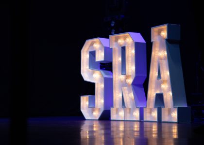 Large illuminated letters spelling S R A, on a stage