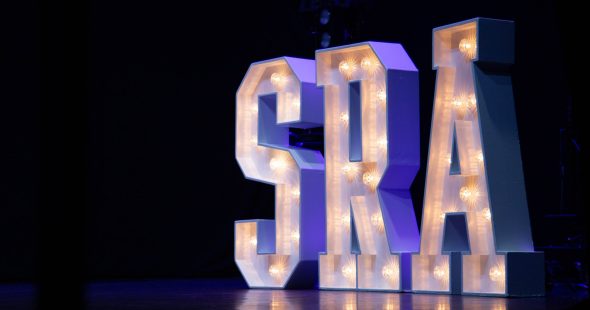 Large illuminated letters spelling S R A, on a stage